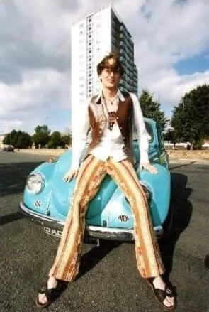 Russell is wearing sandals, flares, and a leather waistcoat while leaning on a Morris Minor bonnet in this promo photograph from the 1990s.