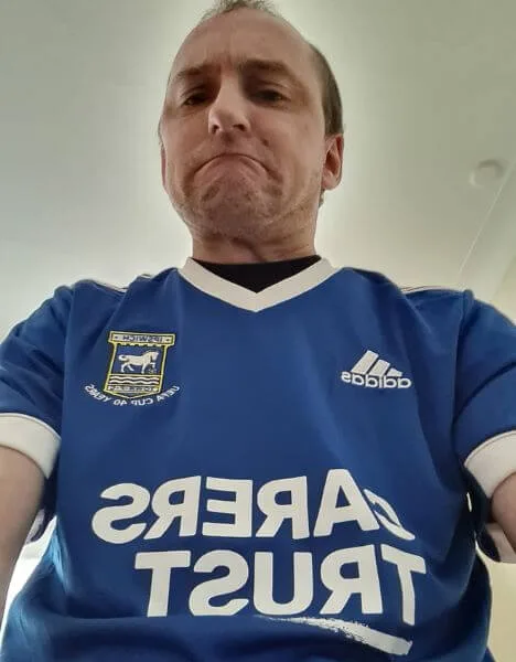 Russell wears an Ipswich Town football shirt with an exaggerated expression of misery.